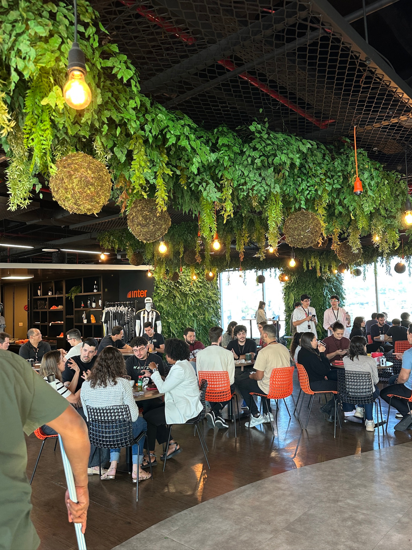 Praça de alimentação do Banco Inter sob o teto verde preservado, com colaboradores nas mesas