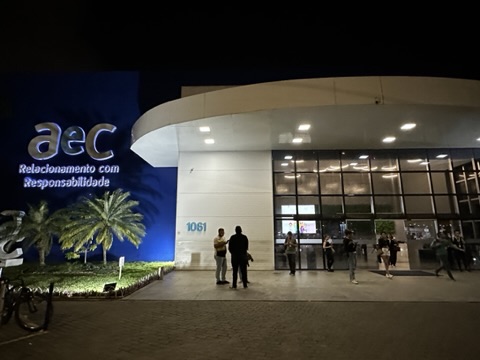 Fachada da unidade da AeC à noite, com marca iluminada e movimento de colaboradores na entrada
