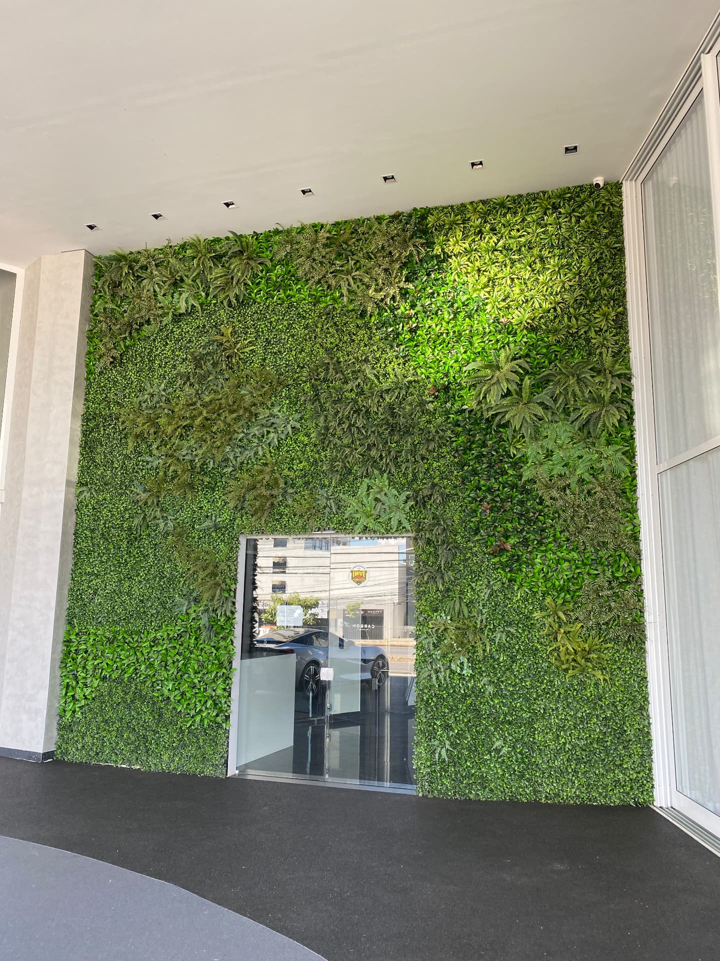 Parede verde permanente da entrada da Avantgarde Motors em plano frontal, com mix de folhagens e porta de vidro ao centro