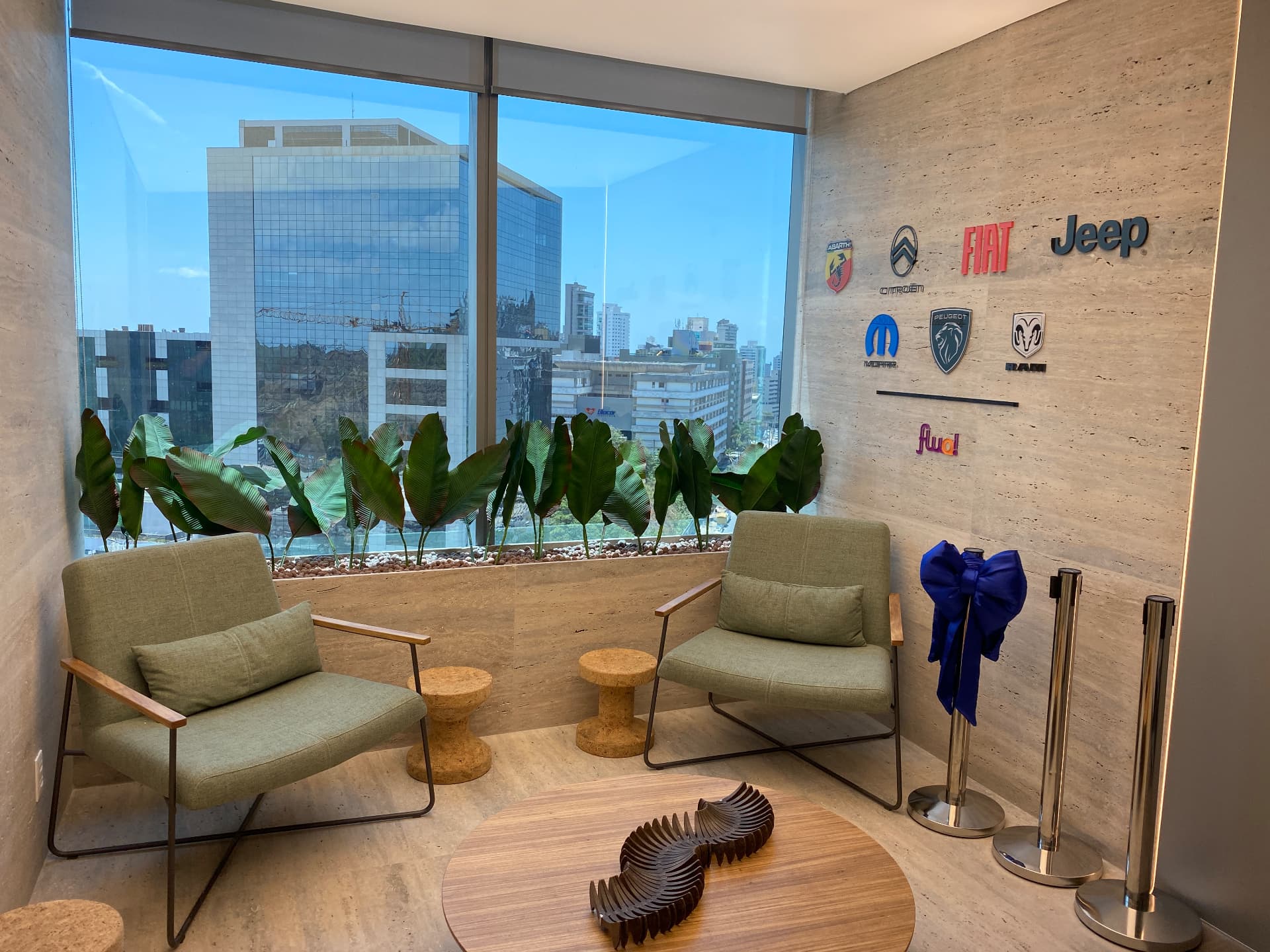 Lounge de espera da Stellantis com bananeiras diante da vista da cidade e parede com as marcas do grupo